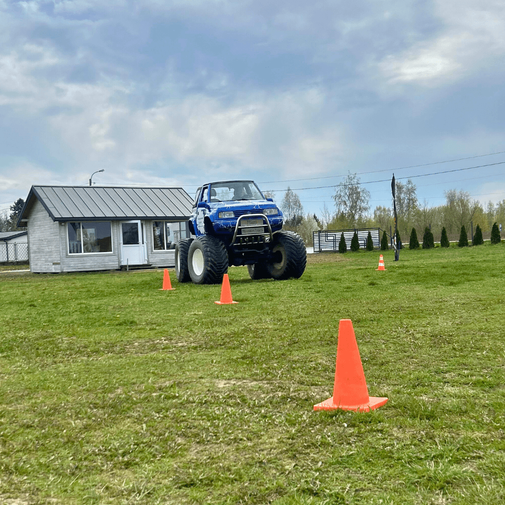 Niebieski monster truck jedzie po trawie wśród pachołków, na tle białego budynku w malowniczej okolicy.