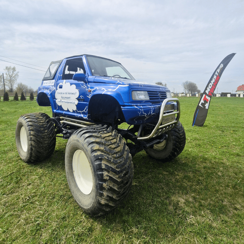 Na zdjęciu widzimy niebieskiego monster trucka stojącego na zielonej trawie, obok flagi promującej atrakcje dla fanów motoryzacji w Gassy k. Warszawy.