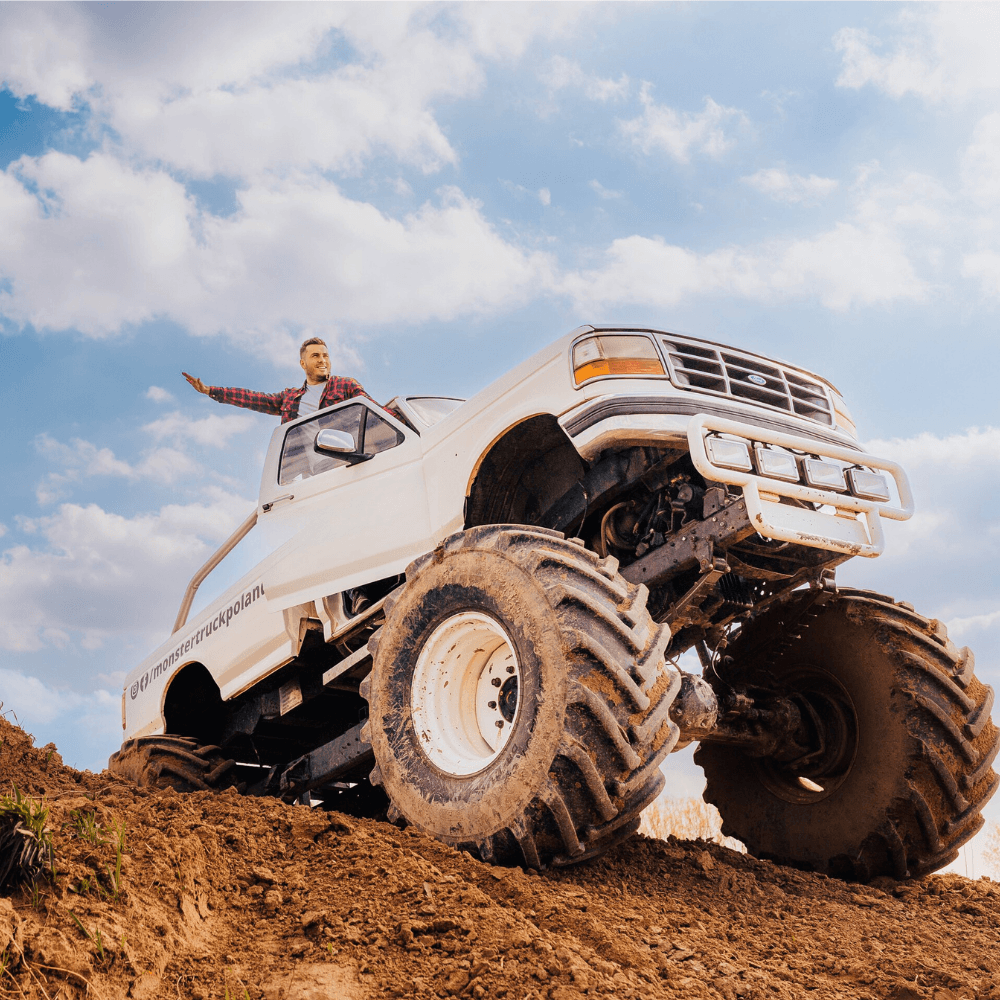 Ogromny monster truck wznosi się na wzgórzu, a z radością wychyla się z niego osoba, podkreślając emocjonującą przygodę, jaką oferuje to wydarzenie w Gassy koło Warszawy.