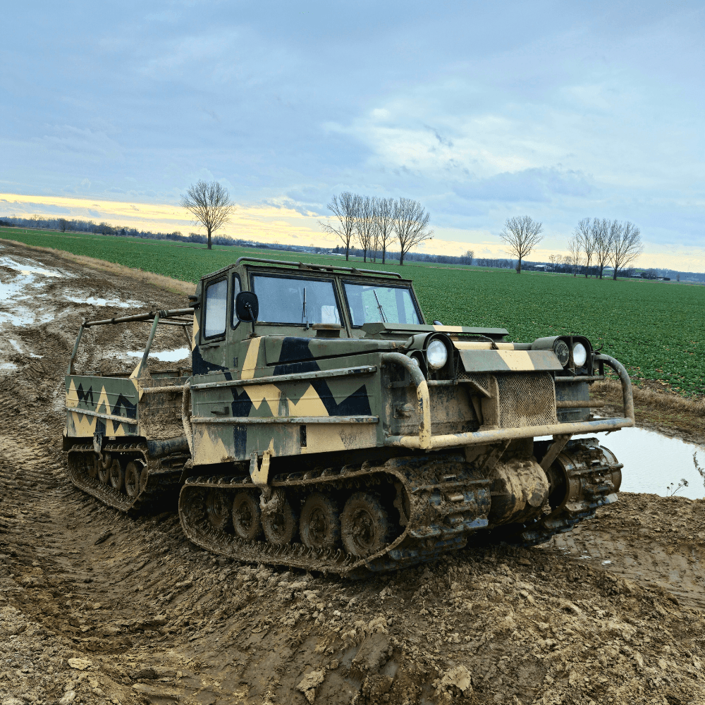 Na zdjęciu widać pojazd gąsienicowy w militarnych barwach, przemierzający błotnisty teren na wiejskich obrzeżach.