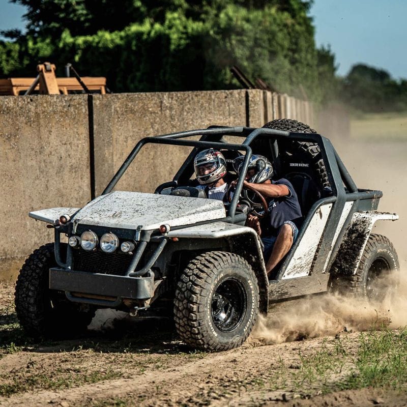 Na grafice widać zacięty wyścig terenowym buggy, którego kierowcy pokonują piaszczystą trasę z dużą prędkością, wzbijając kurz w powietrze.