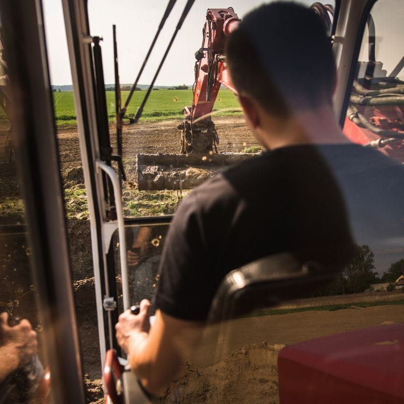 Zdjęcie przedstawia osobę obsługującą koparkę w otoczeniu rozległych pól, co może być jedną z atrakcji związanych z monster truckami w Gassy pod Warszawą.