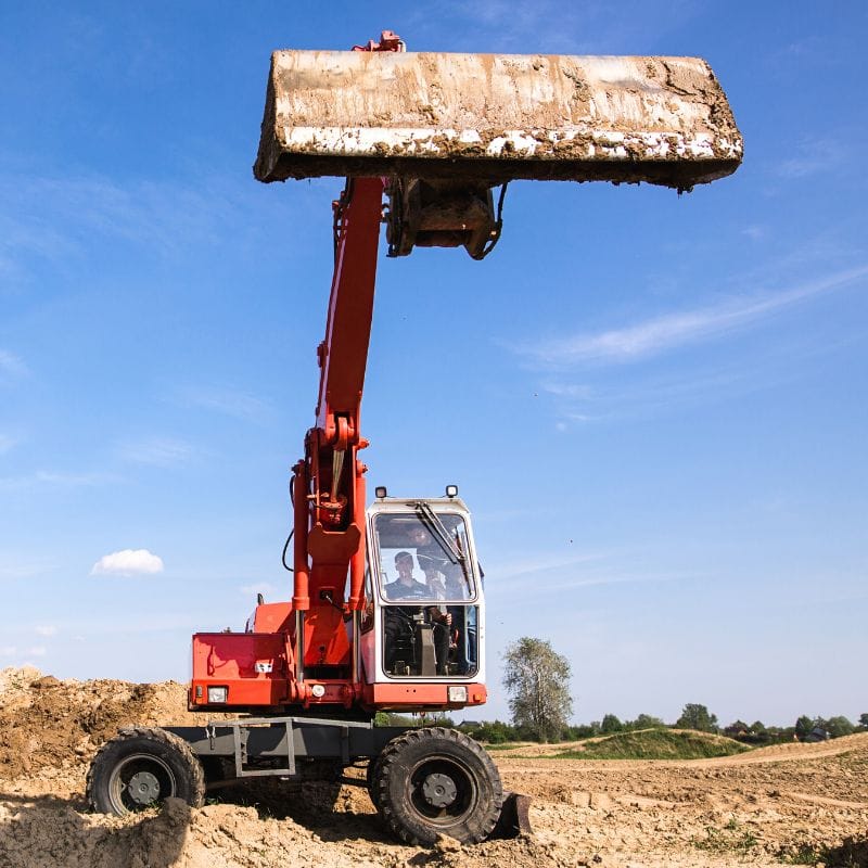 Na fotografii widzimy czerwoną koparkę manewrującą na piaszczystym terenie, co może być częścią przygotowań przed atrakcjami z monster truckami w Gassy koło Warszawy.