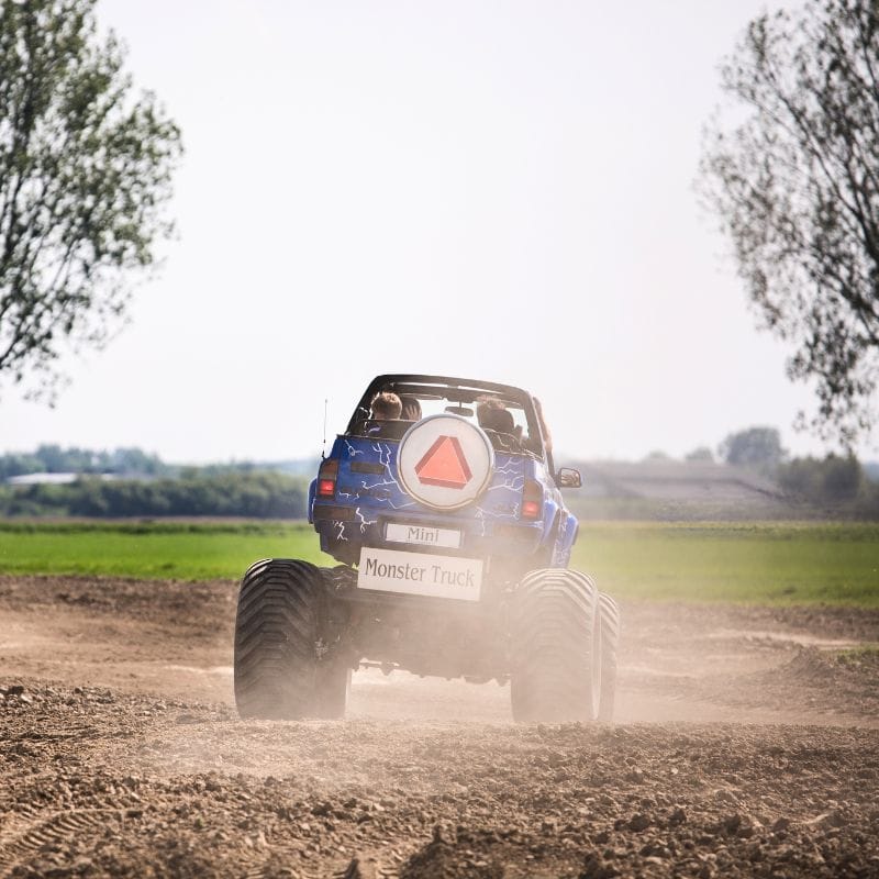 Na obrazie widać mini monster trucka podczas jazdy po piaszczystej trasie na łonie natury, zadowolonych pasażerów i widok na zielony pejzaż w tle.