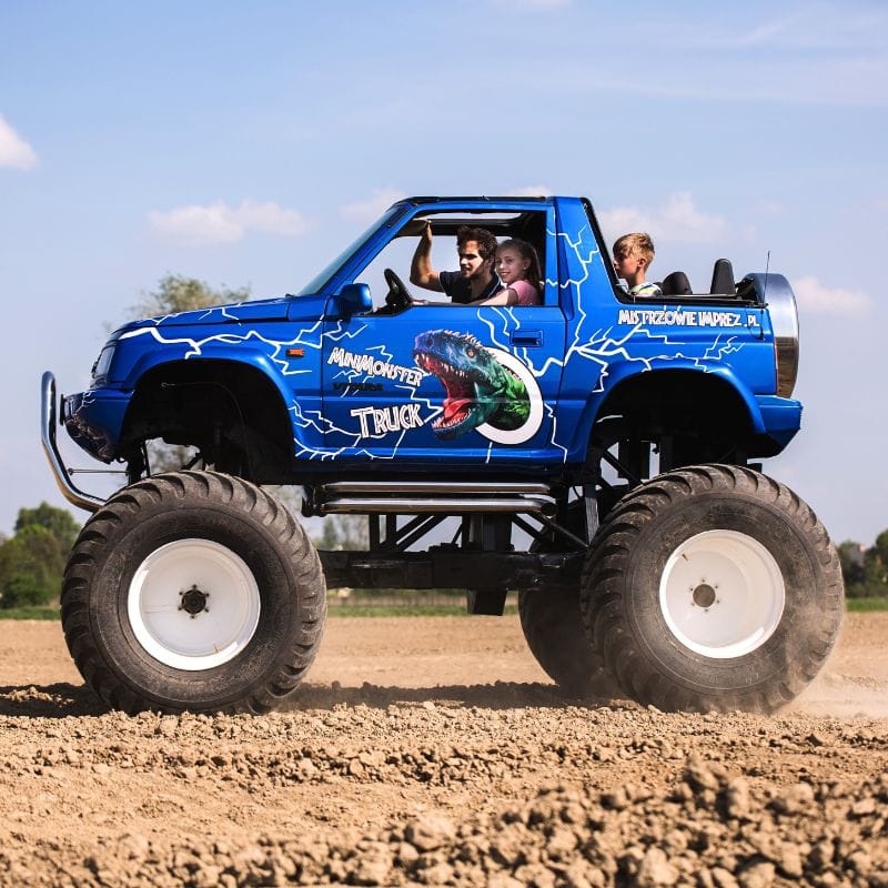Na zdjęciu widać niebieskiego monster trucka z pasjonatami w środku, przemierzającego teren, z logo MiniMonster Truck na boku.