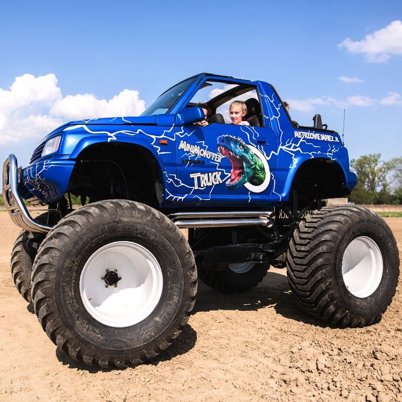 Na zdjęciu widać niebieski monster truck z dużymi kołami, ozdobiony grafiką dinozaura, jadący po piaszczystej nawierzchni, z uśmiechniętym dzieckiem za kierownicą.