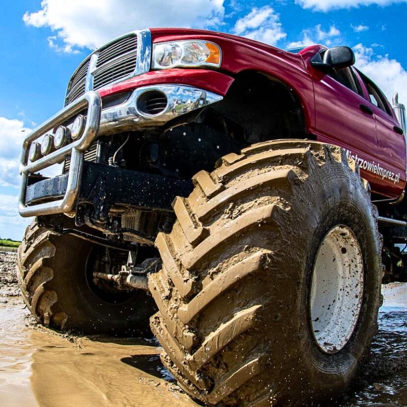 Czerwony monster truck z błotnistymi kołami radzi sobie z terenem pod błękitnym niebem, podkreślając emocje związane z atrakcjami terenowymi dla wszystkich grup wiekowych w pobliżu Warszawy.