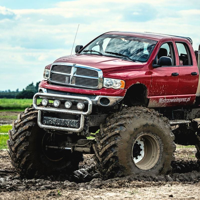 Na grafice przedstawiono imponujący, czerwony monster truck przemierzający błotnisty teren, symbolizujący atrakcje dla fanów tego typu pojazdów w Gassy pod Warszawą.