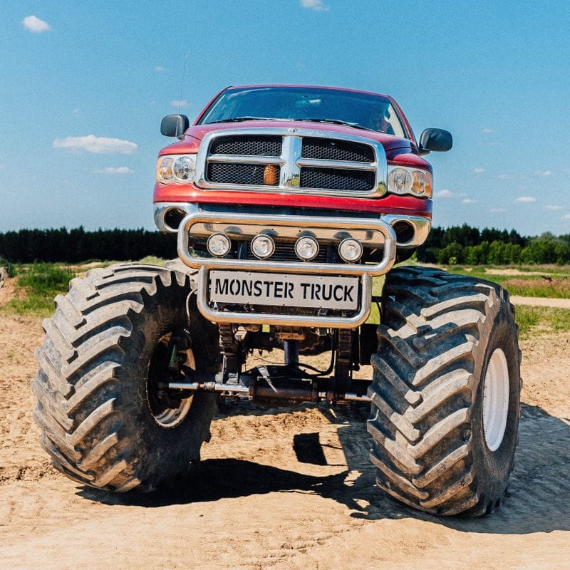 Czerwona ciężarówka typu monster truck, stojąca na dużych terenowych oponach, dominuje na piaszczystym terenie w słoneczny dzień.