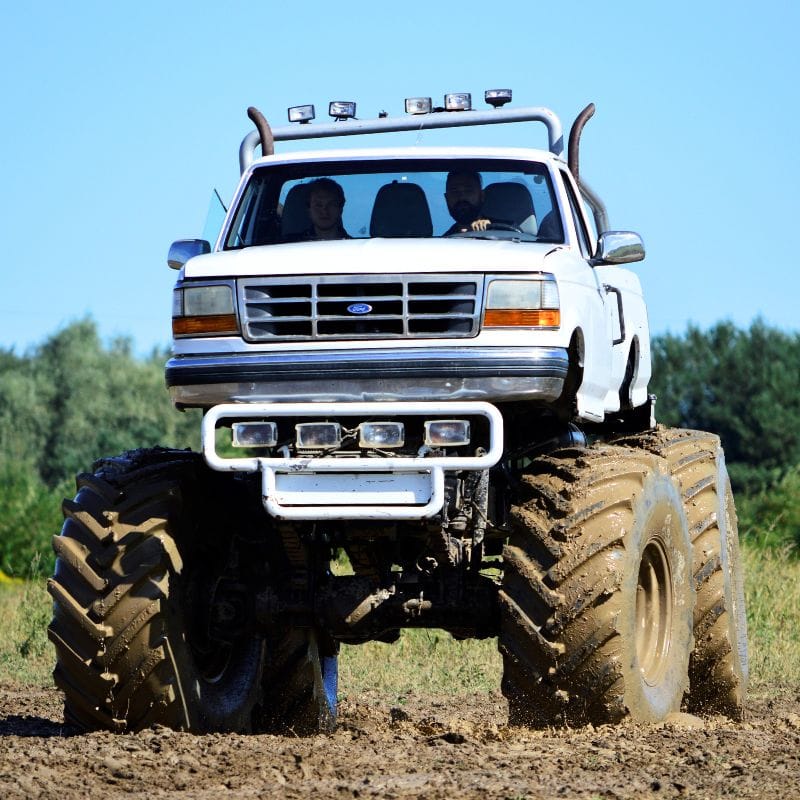 Imponujący biały monster truck z masywnymi oponami jedzie po błotnistej drodze, zapewniając ekstremalne przeżycia dla wszystkich fanów adrenaliny.
