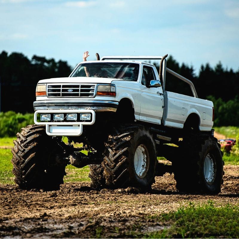 Ogromny biały monster truck przejeżdża przez błotnisty teren, a zza okna widać rękę machającą w geście radości.