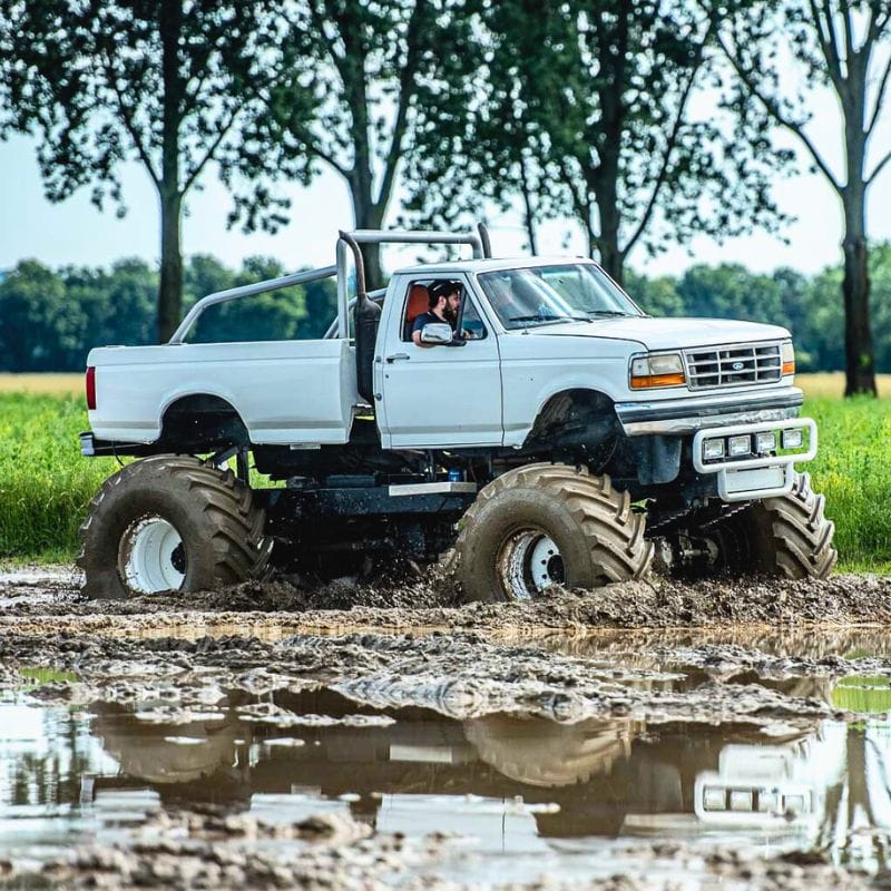 Biały monster truck przejeżdża przez błotnisty teren, co zapewnia ekscytującą przygodę dla pasażerów i widzów.