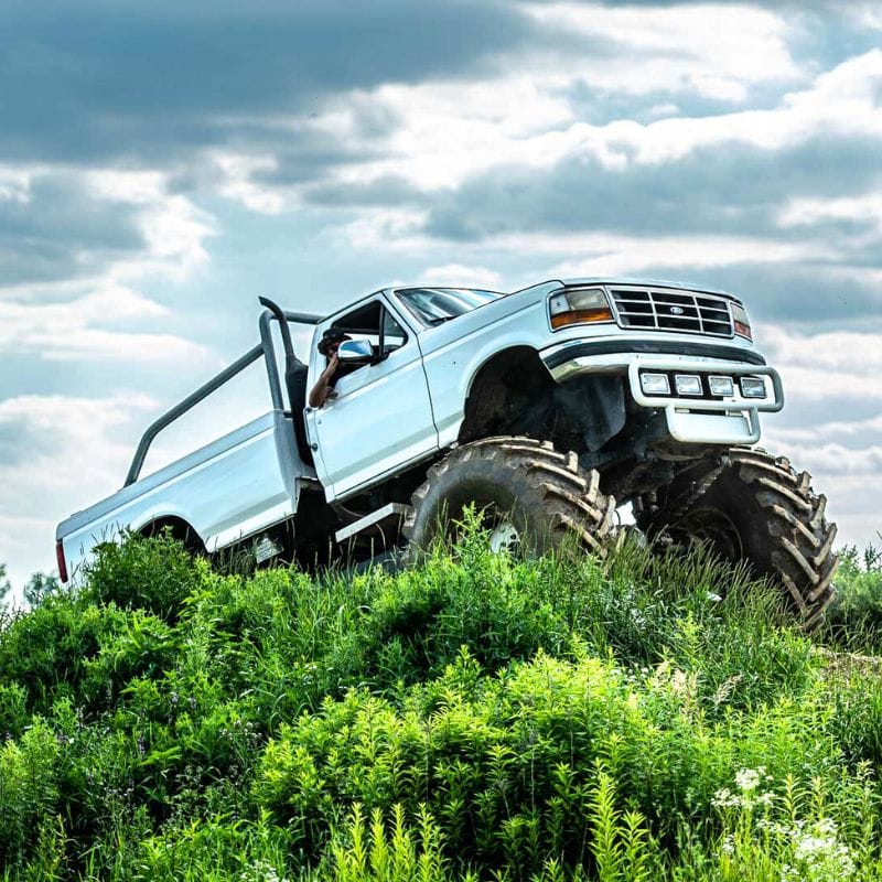 Potężny, biały monster truck z dużymi oponami wjeżdża na zielone wzniesienie pod zachmurzonym niebem.