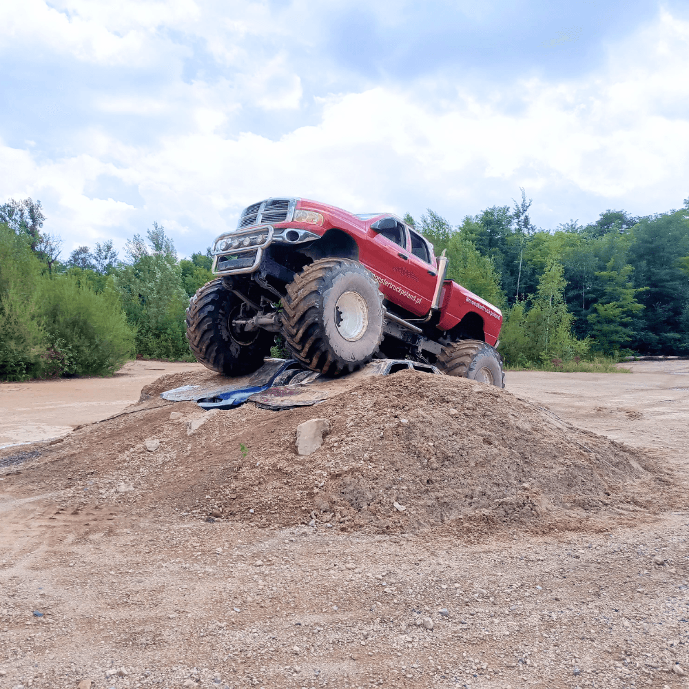 Na zdjęciu potężny monster truck w intensywnym czerwonym kolorze triumfalnie wznosi się na piaszczystym wzniesieniu, otoczony soczystą zielenią lasu w pobliżu siedziby w Gassy koło Warszawy.
