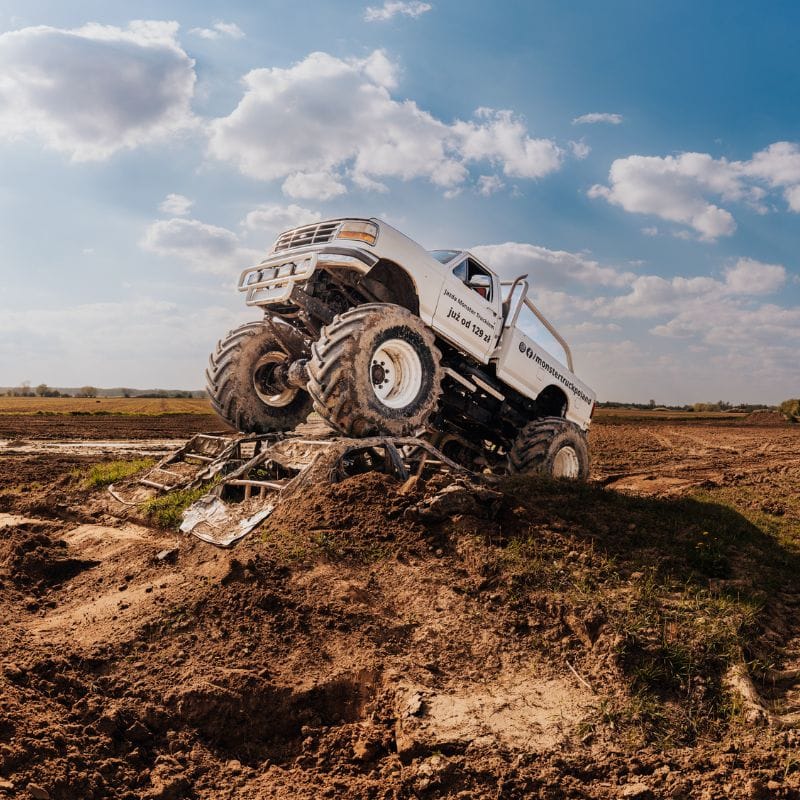 Białe monster trucki z wielkimi oponami pokonują terenowe przeszkody na torze pod błękitnym niebem, oferując emocje dla dorosłych i dzieci w pobliżu Gassy pod Warszawą.