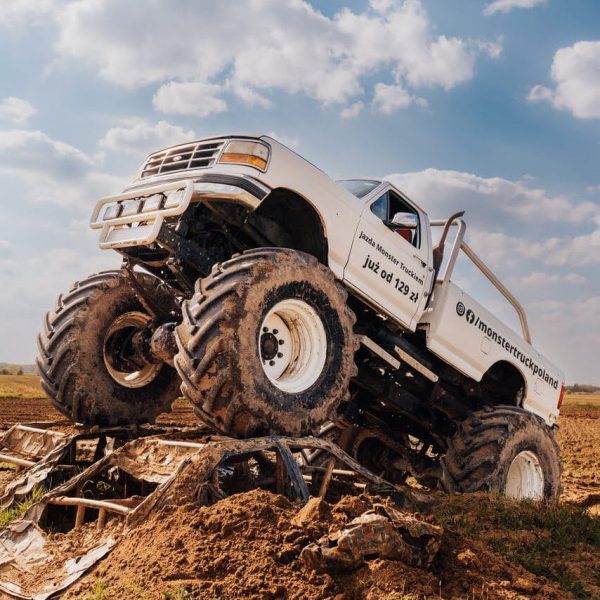 Na zdjęciu przedstawiony jest potężny monster truck w akcji, wspinający się na wrak samochodu na błotnistej trasie, pokazujący swoją siłę i atrakcyjność dla entuzjastów motoryzacji w Gassy pod Warszawą.