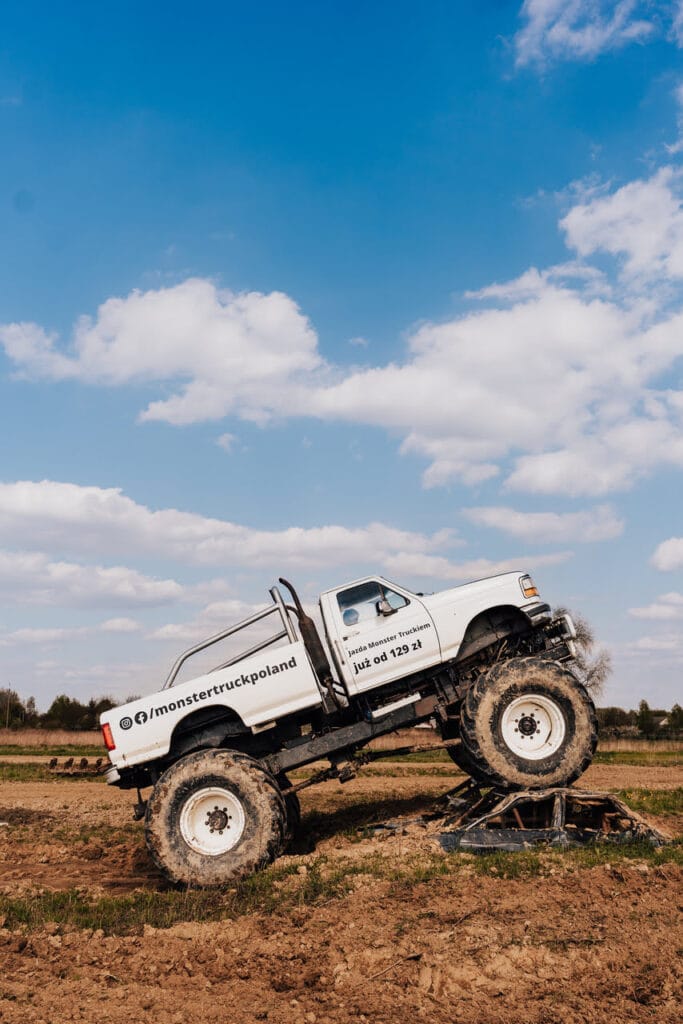 Na zdjęciu widzimy monster trucka wspinającego się na wrak samochodu na piaszczystym terenie pod bezchmurnym niebem, co sugeruje emocjonującą atrakcję, idealną dla fanów mocnych wrażeń w pobliżu Warszawy.