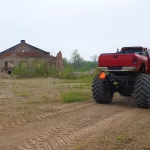 Czerwony monster truck z potężnymi kołami stoi na rzadko zarośniętym terenie, a w tle widać opuszczony budynek z czerwonej cegły.