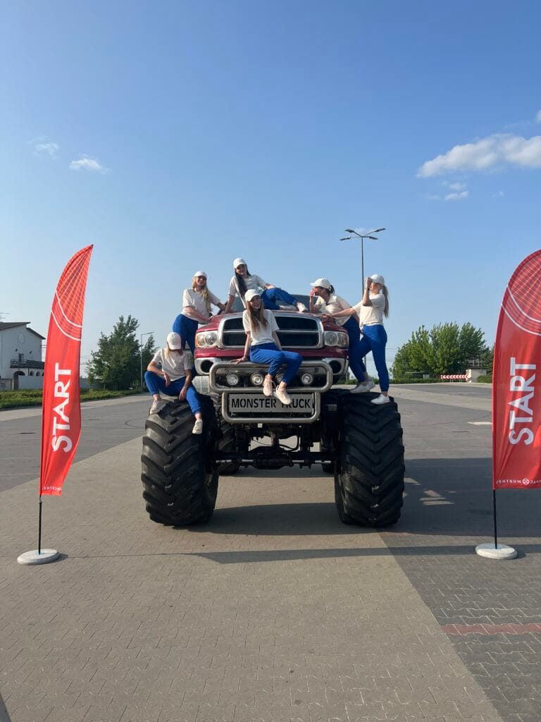 Na zdjęciu widać grupę osób pozujących na ogromnym monster trucku otoczonym dwoma czerwonymi flagami z napisem START.