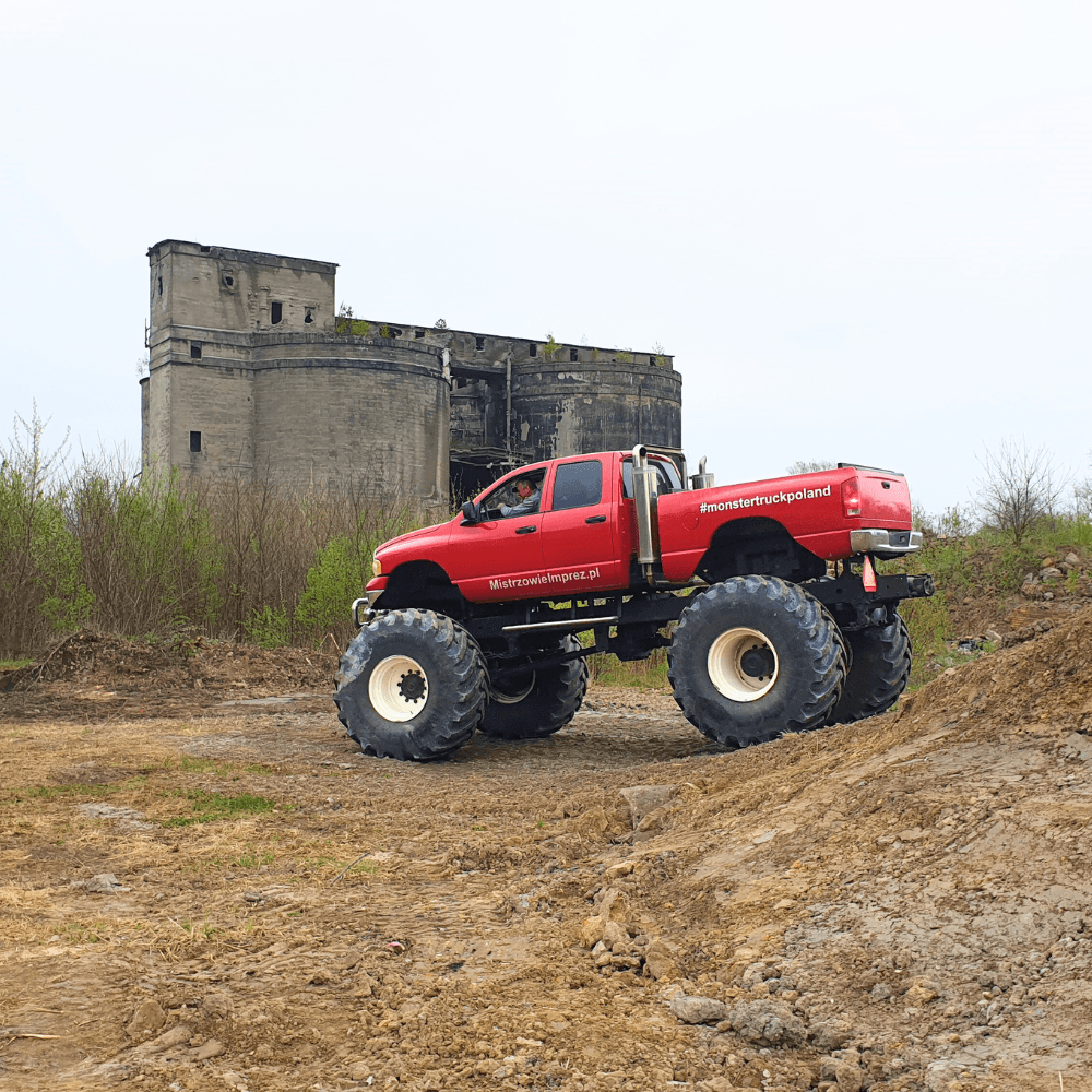 Gigantyczny monster truck w intensywnym czerwonym kolorze stoi na nierównej, ziemistej nawierzchni, a w tle widać zrujnowany, imponujący budynek, tworząc widowiskową scenerię idealną dla miłośników ekstremalnych atrakcji.