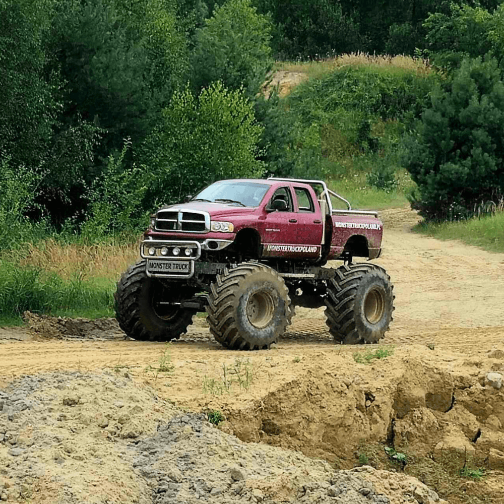 Na zdjęciu widać ogromny monster truck w czerwonym kolorze przemierzający piaszczysty teren otoczony zielenią.