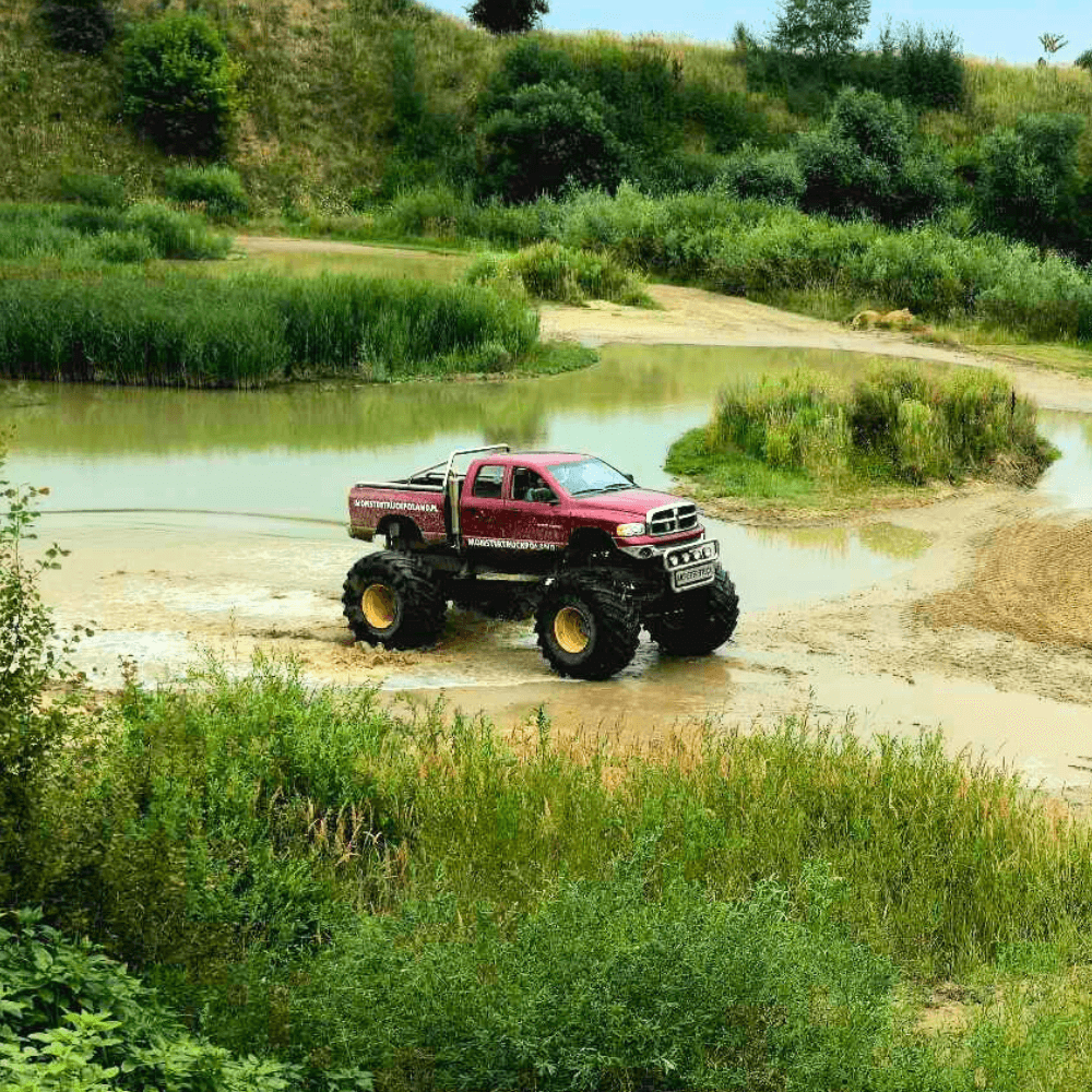 Monster truck w intensywnym czerwonym kolorze przemierza błotnisty teren otoczony bujną zielenią, idealnie oddając emocje i przygody, jakie czekają na miłośników dużych maszyn w Gassach pod Warszawą.