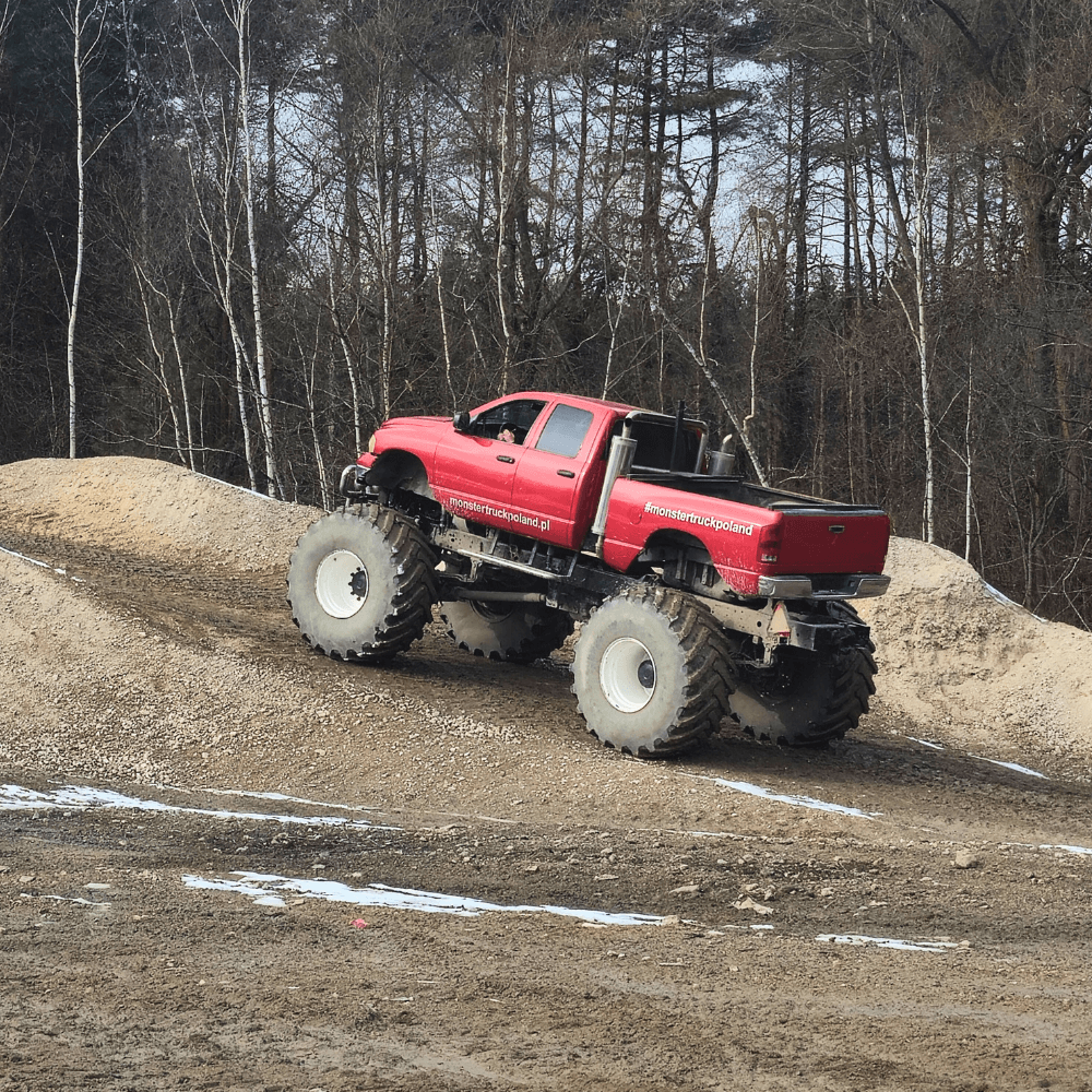 Na grafice czerwony monster truck z potężnymi kołami przemierza piaszczysty teren, oferując ekscytujące przeżycia terenowe w okolicach Gassy pod Warszawą.
