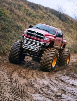 Czerwony monster truck z ogromnymi kołami przemierza błotnistą trasę w malowniczym otoczeniu.