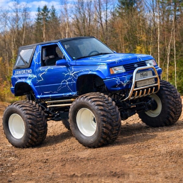 Niebieski mini monster truck z błyskawicznym motywem graficznym prezentuje się imponująco na piaszczystym terenie, stanowiąc atrakcję dla miłośników ekstremalnych doznań w okolicach Warszawy.